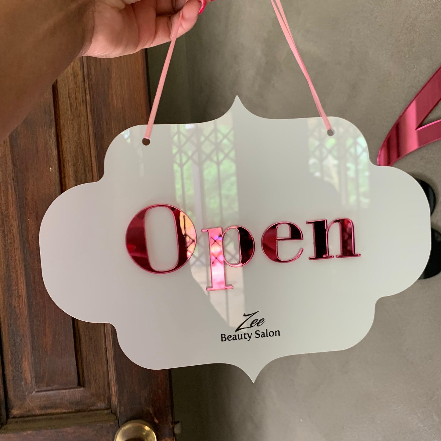 Hand holding a 'Open' sign for Zee Beauty Salon against a door background