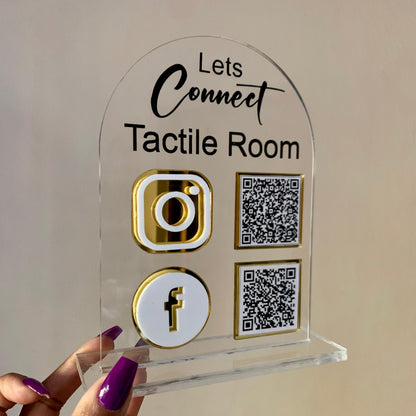 Clear acrylic sign with social media icons and QR codes held by a hand against a neutral background
