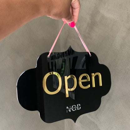 Hand holding a 'Open' sign with gold text on a gray background